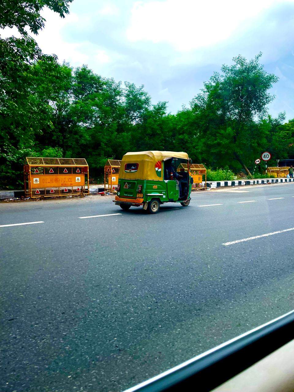 A green and yellow auto rickshaw driving along a tree-lined road in Delhi, seen through a car window.