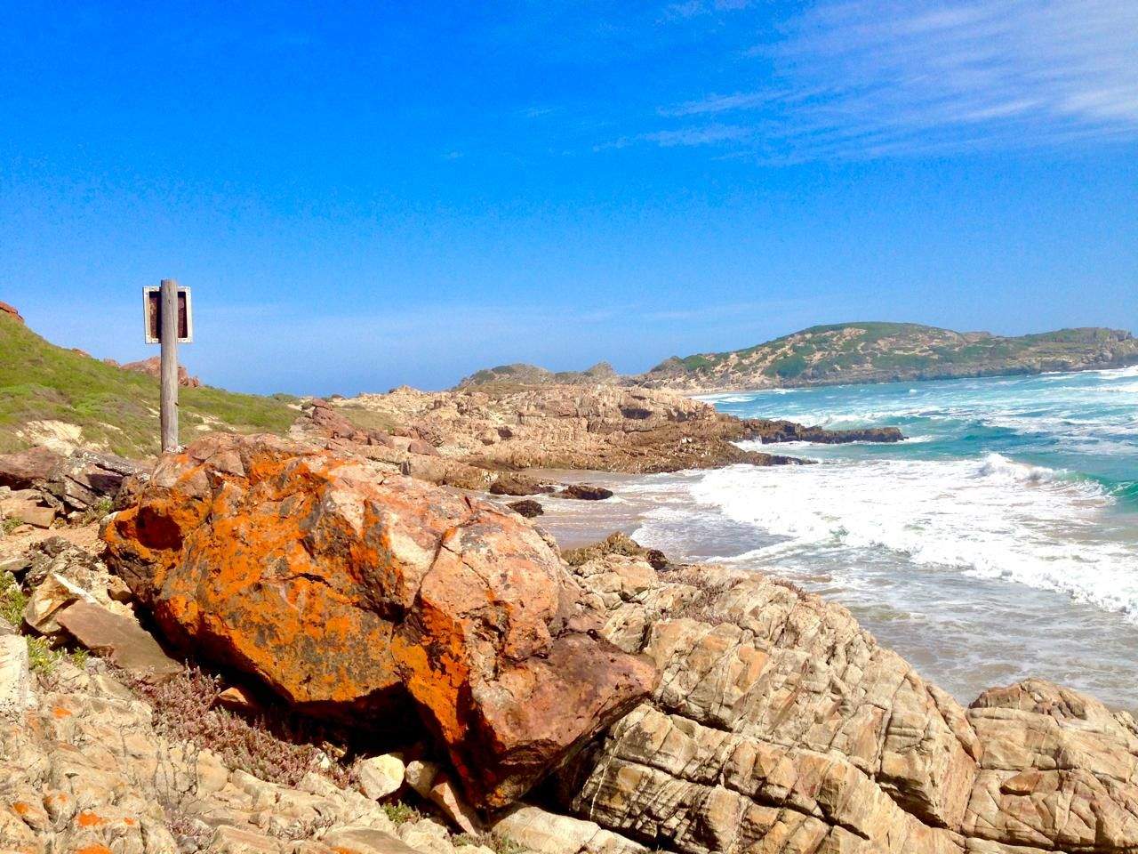 Waves breaking against a rocky coastline at Robberg Nature Reserve, with a trail marker post in the foreground.